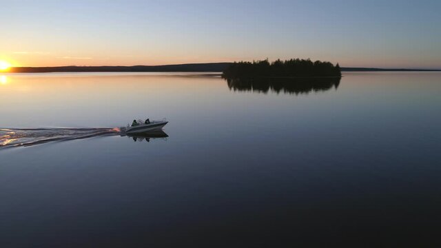 Aerial, tracking, drone shot, of a boat, driving in calm, reflecting waters, of lake Posionjarvi, midnight sun, on midsummer evening, in Posio, Lapland, Finland