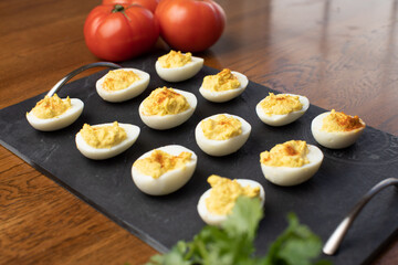 Photo of a tray full of devilled eggs on a wooden table. There is tomatoes also on the table.