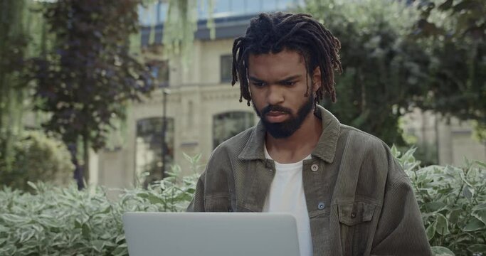 Crop view of young concentrated guy with dreadlocks working on laptop while sitting in park . Bearded serious afro american male student in casual clothes styding outdoors