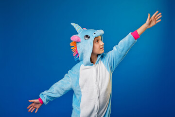 Charming girl playfully poses in the costume of the unicorn. A Studio shot of an emotional woman in kigurumi having fun on a blue background. Cheerfully spreading his hands to the sides.