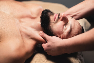 Obraz premium Male hands of a masseur doing neck massage to a bearded male athlete in a dark room of the massage room
