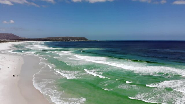 Scenic drone footage of breaking waves, ocean, wide white sand beach in Noordhoek Beach in Western Cape in South Africa. Cliffs and mountains in the background. Drone flying above turquoise water.
