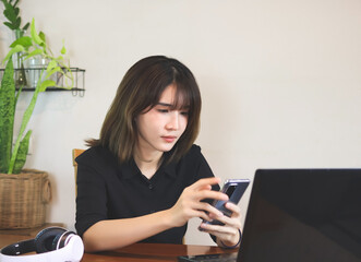 Asian woman wearing black shirt, sitting at wooden table with computer laptop and headphones, using mobile phone. white wall with house plant background.