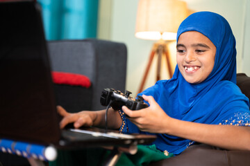 Fototapeta na wymiar Happy smiling Muslim Girl Kid playing online videogame on laptop by using gamepad or joystick at home by wearing hijab dress - concept of kid using technology and modern lifestyle.