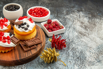 bottom view berry cakes and chocolates on chopping board bowls with pomegranate raisins and raspberries and colorful pinecones on grey-white background with copy space