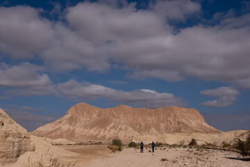 Fototapeta na wymiar Two male friends on a hiking trail in a remote desert region. Panoramic landscape of dry wide wadi with colorful sandy hills and mountain folds . White clouds on the blue sky in a winter sunny day.