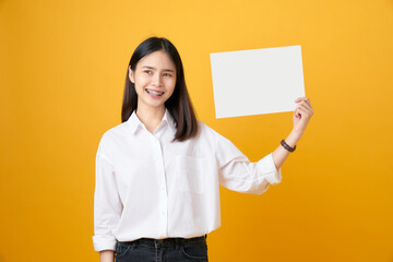 Fototapeta na wymiar Young Asian woman holding blank paper with smiling face and looking on the yellow background. for advertising signs.