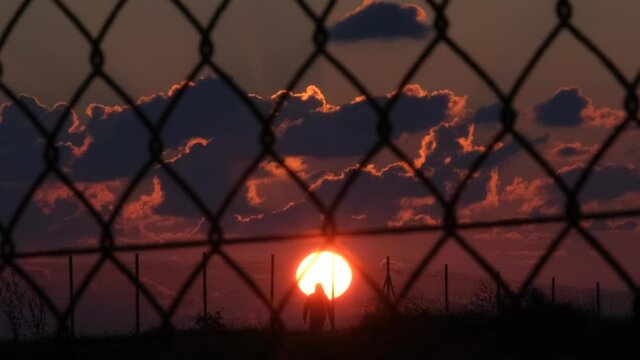 Mysterious point of view from behind a fence, watching a person walking alone down a nature path under the evening sunset.