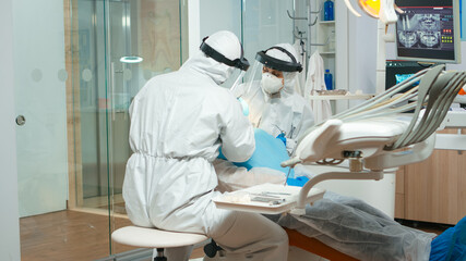 Dentist with medical overall checking senior patient teeth problem during coronavirus pandemic. Doctor and nurse working wearing coverall, face shield, protection suit, mask, gloves in dental office