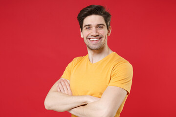 Obraz premium Young smiling unshaved cheerful friendly caucasian handsome man 20s wearing casual basic yellow t-shirt looking camera holding hands crossed folded isolated on red color background studio portrait.