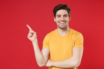 Young smiling unshaved caucasian handsome cheerful man 20s in casual yellow t-shirt pointing index finger aside on workspace copy space area mock up isolated on red background studio portrait.