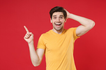 Young surprised unshaved man in casual blank print design yellow t-shirt pointing index finger aside on workspace copy space area mock up put hand on head isolated on red background studio portrait.