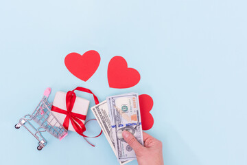 A hand holds cash dollars over a shopping cart with a gift box and red love hearts on a blue background, copy space