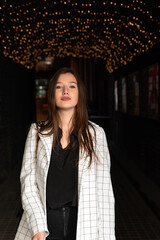 Fototapeta premium Portrait of brunette girl on evening street and garlands background. Vertical frame