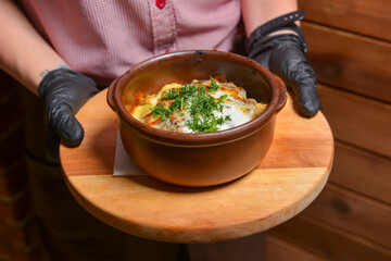 Roasted vegetables and meat under melted cheese in a pot. Baked potatoes dish. Waiter serving dinner at restaurant.