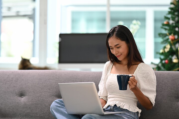 Cheerful young woman using laptop watching funny video and drinking coffee while sitting on couch in living room.