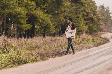 A slender woman of model appearance with long dark hair is dressed in a fur coat, a sweater hat and leather pants walks along a path against a background of a pine forest in winter