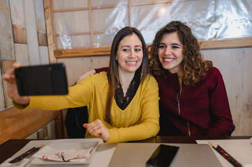 Two beautiful young cheerful women taking a selfie photo in a restaurant hugging with smiling faces. Friendship, togetherness concept.