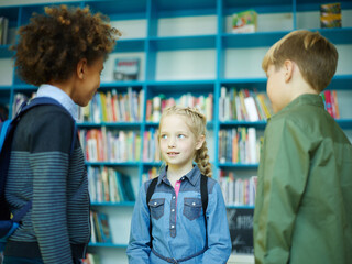 Group of three diverse elementary students communicating friendly near book shelves in school library