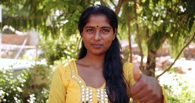 Slow-motion close-up portrait of a young adult Indian woman outdoors raises one hand thumbs up for best of luck wishes at camera pov- defocused racking focus blurred 