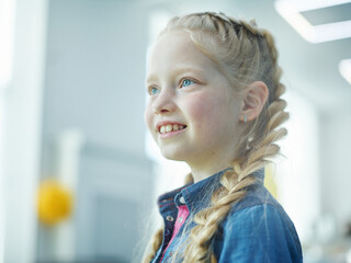 Cute little girl with braided hair and freckles standing in school lobby and looking away dreamily...
