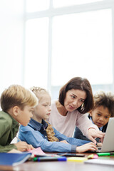 Female teacher explaining computer science to group of curious diverse elementary students looking at laptop screen sitting at desk in school classroom