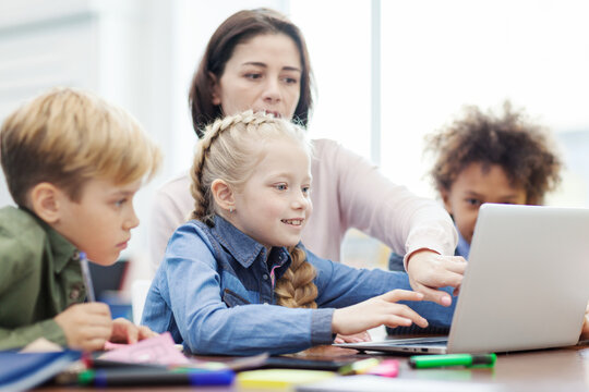 Female teacher explaining computer science to group of diverse elementary students looking at laptop screen sitting at desk in classroom