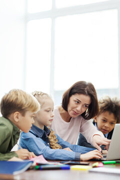 Female Teacher Explaining Computer Science To Group Of Curious Diverse Elementary Students Looking At Laptop Screen Sitting At Desk In School Classroom