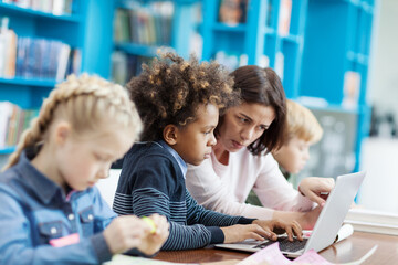 Side view selective focus of mixed race elementary student doing task on laptop computer helped by female teacher, his defocused classmates studying sitting at desk in school classroom nearby