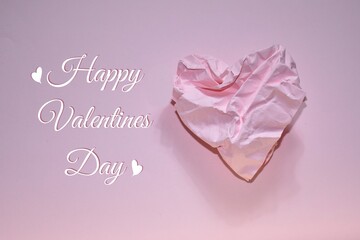 Pastel pink abstract background with the words "Happy Valentines Day" and heart made of crumpled paper. Crumpled paper in the shape of heart on pink background. Space for text. Valentine