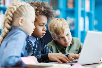 Side view of three curious diverse students using laptop sitting at desk in school library. Mixed ...