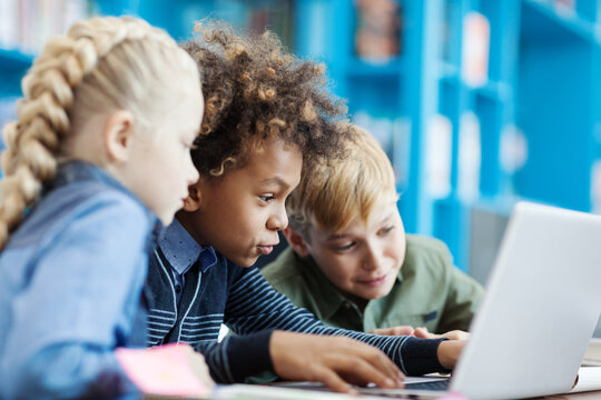 Group of three excited diverse students using laptop sitting at desk in school library. Mixed race schoolboy and his friends browsing through information on Internet