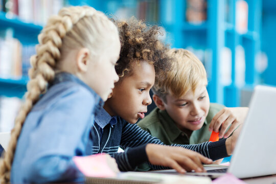 Group of three curious diverse kids using laptop sitting at desk in school library. Mixed race schoolboy and his friends browsing through information on Internet