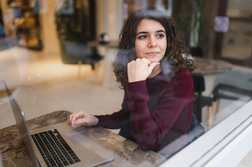 Close-up young woman portrait sitting in a cafe with chin in hand and relaxed face looking out the window. Reflections on the glass. Leisure activities, relaxation concept.