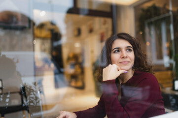 Close-up young woman portrait sitting in a cafe with chin in hand and relaxed face looking out the window. Reflections on the glass. Leisure activities, relaxation concept.