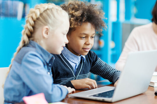 Two elementary school kids, mixed race boy and Caucasian girl, studying on laptop computer sitting at desk in classroom