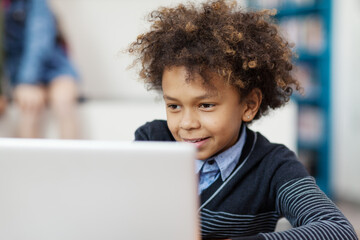 Mixed race curly-haired schoolboy browsing Internet on laptop computer in classroom and smiling