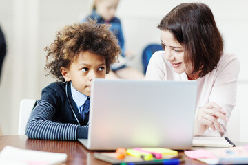 Smiling female teacher explaining thoughtful mixed race boy task on laptop computer sitting at desk in elementary school classroom