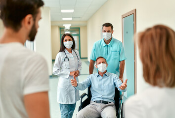 Obraz premium A nurse and a young doctor wearing surgical masks to protect against the covid 19 pandemic take care of a mature male patient in a wheelchair at a hospital who has been visited by a family.