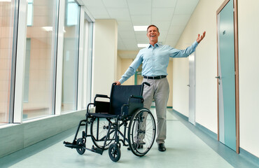 A handsome mature man stands recovered from his disability near a wheelchair and raised his hand in a sign of victory, standing in the clinic corridor.