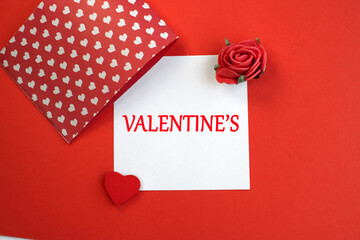 Valentine's day is written on a white sheet which is on a red background with a red heart and a rose. 