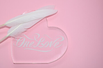 White feathers on transparent glass heart, on pink background. The inscription 