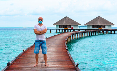a young man wearing a protective face mask stands on a pier near water villas in the Maldives, a...