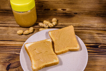 Glass jar with peanut butter and plate with sandwiches on a wooden table