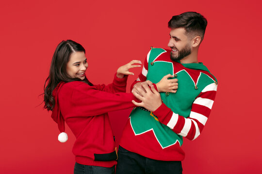 Positive Couple Dressed In Christmas Funny Sweaters. Woman Tickling Her Boyfriend Over Red Background.