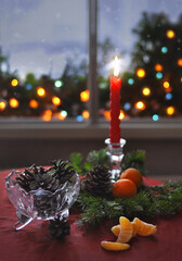 A red candle, fir branches, tangerines and cones on the table near a snowy window. Lights outside the window.