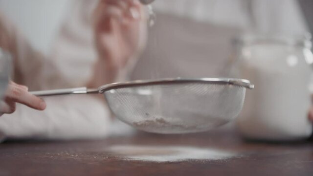 Midsection slow-motion close-up of unrecognizable female hands sifting white flour at kitchen table using sieve and tablespoon