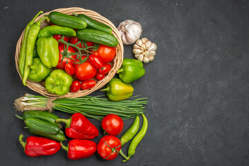 Fresh vegetables red tomatoes with stems green peppers and cucumbers inside and outside of basket on dark background