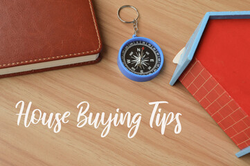 Top angle view of toy wooden house, compass and notebook written with text HOUSE BUYING TIPS....