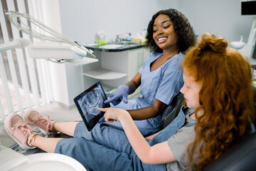 Little patient kid girl with curly red hair sitting in dentistry chair and pointing on the tablet with teeth panoramic radiography. Pretty african woman doctor explaining the ways of treatment